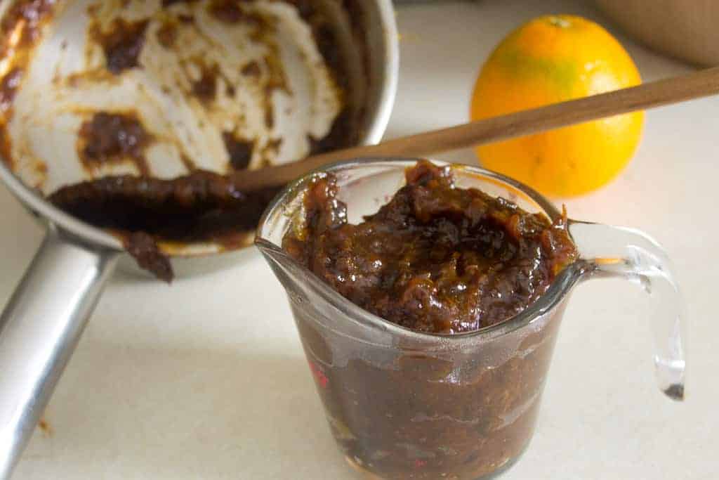 prune filling A glass measuring cup brims with a thick, dark brown prune paste. Behind it, a saucepan holds remnants of the same mixture, hinting at freshly made vegan hamantaschen. An orange and a wooden spoon rest on the counter beside the saucepan.