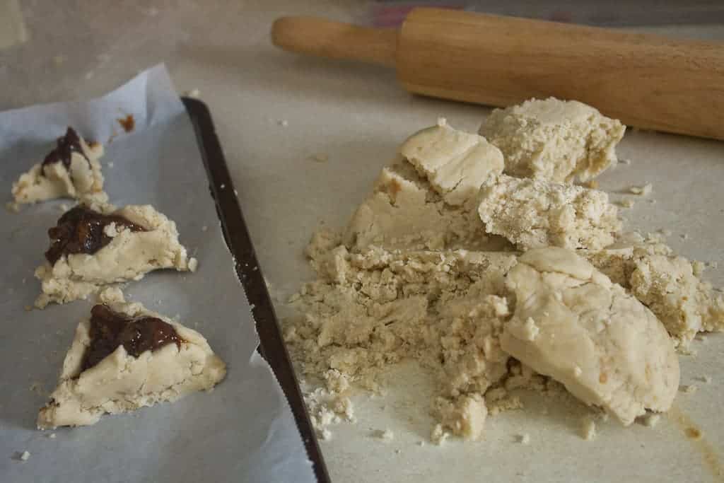 working the dough Unbaked triangular pastries filled with a prune filling rest on a parchment-lined baking sheet. A pile of dough and more unshaped vegan hamantaschen pieces are on the countertop beside a wooden rolling pin.