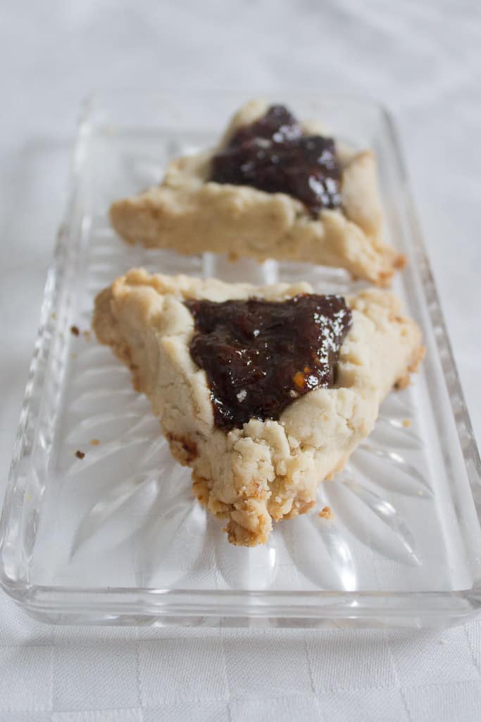 two for tea Two triangular pastries with a crumbly beige crust and dark prune filling, these vegan hamantaschen rest on a clear glass dish adorned with floral patterns. The elegant dish is set on a white, textured surface.