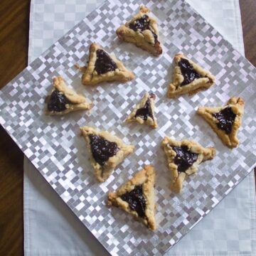 A square silver tray on a white cloth displays eight triangular prune hamantaschen with dark filling. The tray rests on a wooden surface.