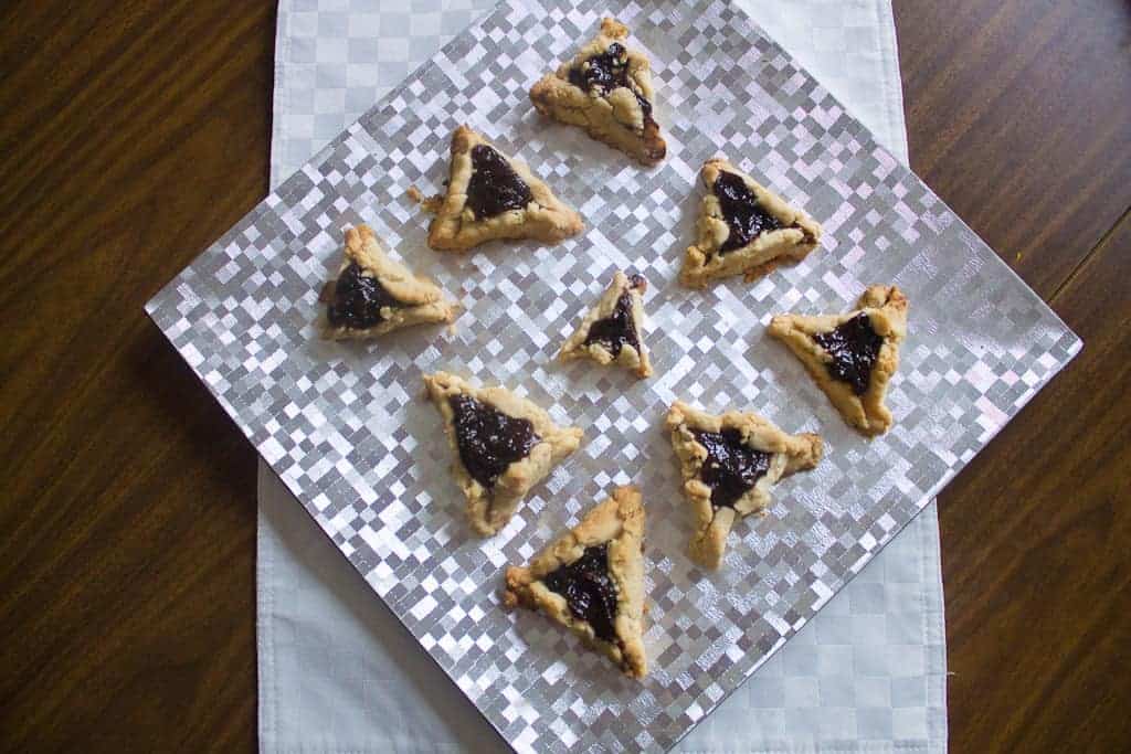 A Purim plate A silver checkered plate holds eight triangular prune hamantaschen filled with dark jam, arranged in a loose circular pattern. The plate rests on a folded white napkin on a wooden table.