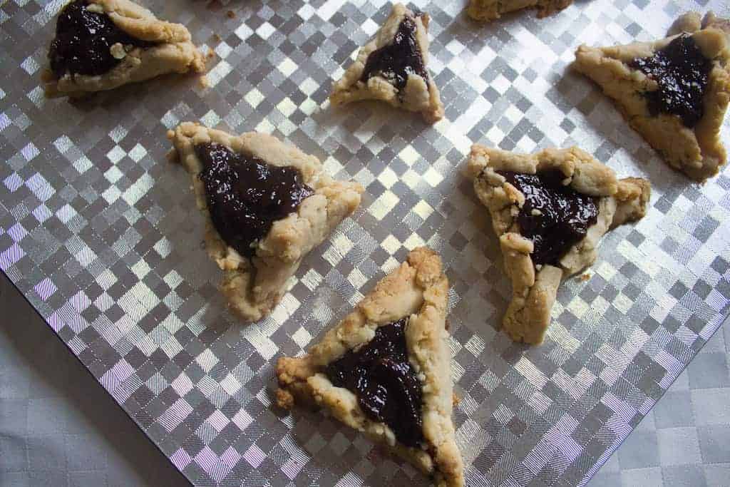 ready to party Hand-crafted prune hamantaschen with a crumbly texture and dark filling are arranged on a reflective, checkered surface. These triangular pastries vary in size with slightly uneven edges, showcasing their artisanal vegan charm.