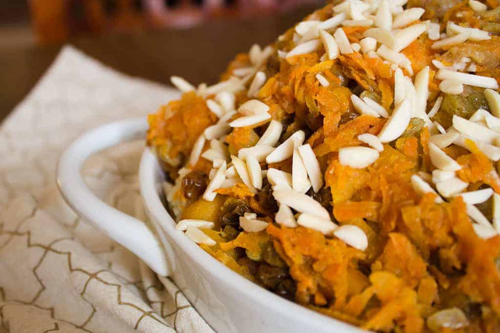 A close-up of a white ceramic dish filled with shredded orange and brown vegetarian pilaf, topped with slivered almonds, resting on a patterned beige napkin.