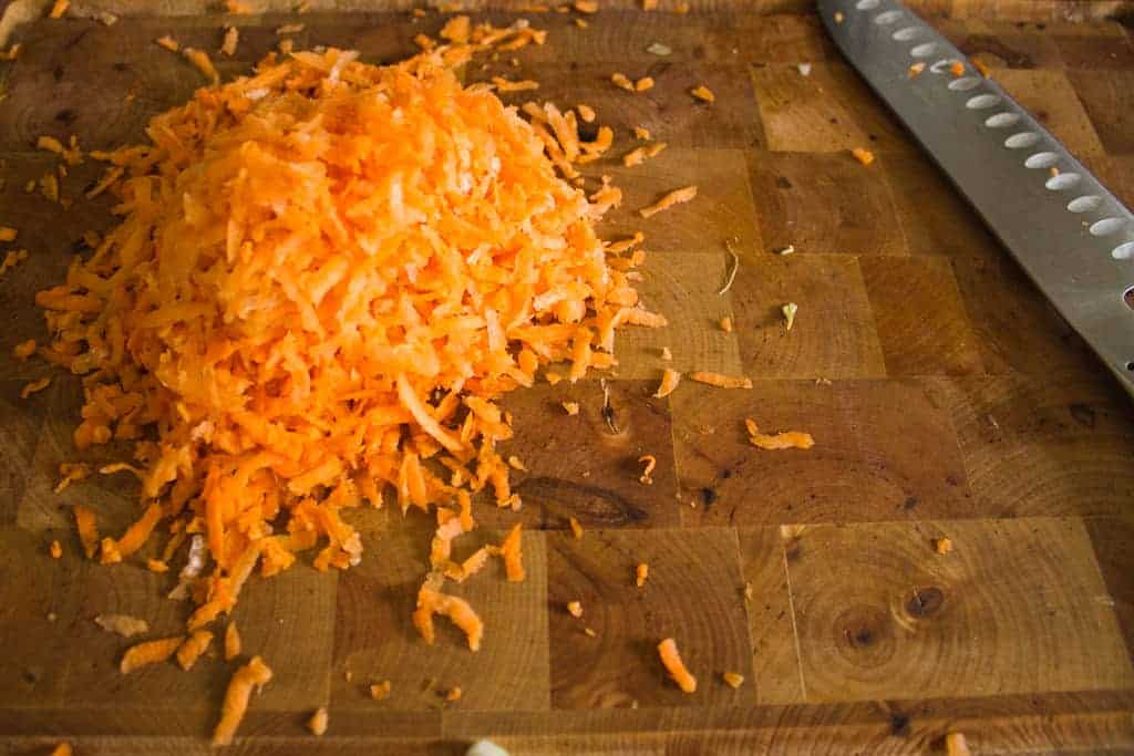 A pile of grated carrots sits on a wooden cutting board next to a knife with oval cutouts, ready to be added to a fresh vegetarian pilaf.