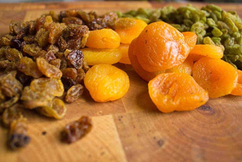 A close-up of dried fruit on a wooden surface, including golden raisins, green raisins, and dried apricots—perfect ingredients for adding sweetness to a vegetarian pilaf—arranged in small piles.