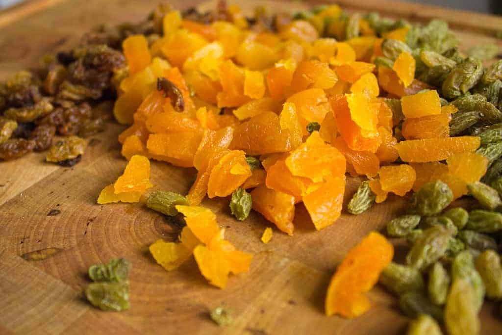 Chopped dried apricots and mixed raisins, perfect for a vegetarian pilaf, are spread out on a wooden cutting board. The apricots are bright orange, while the raisins vary in shades of green and brown.