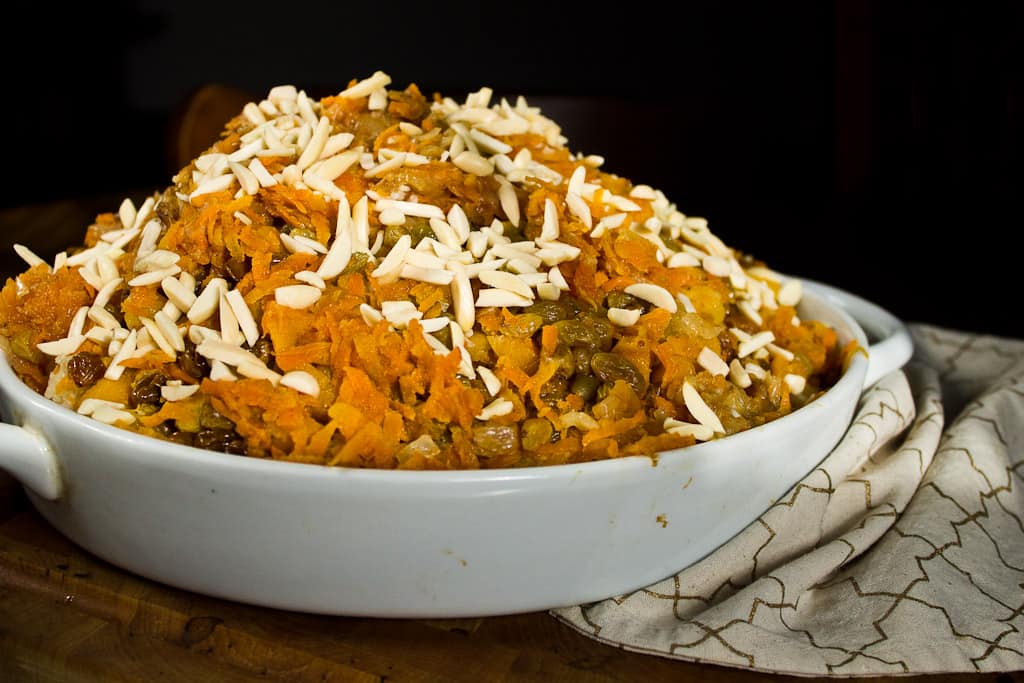 A white dish filled with a mound of orange-colored carrot halwa, topped with slivered almonds, sits on a patterned cloth with a dark background—perfect alongside vegetarian pilaf for an elegant meal.