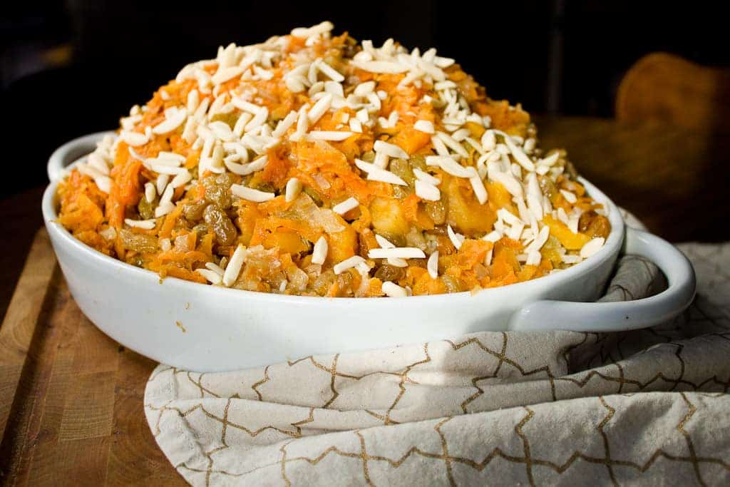 A white oval baking dish filled with a layered vegetarian pilaf casserole topped with shredded cheese, sitting on a beige and white patterned cloth on a wooden surface.