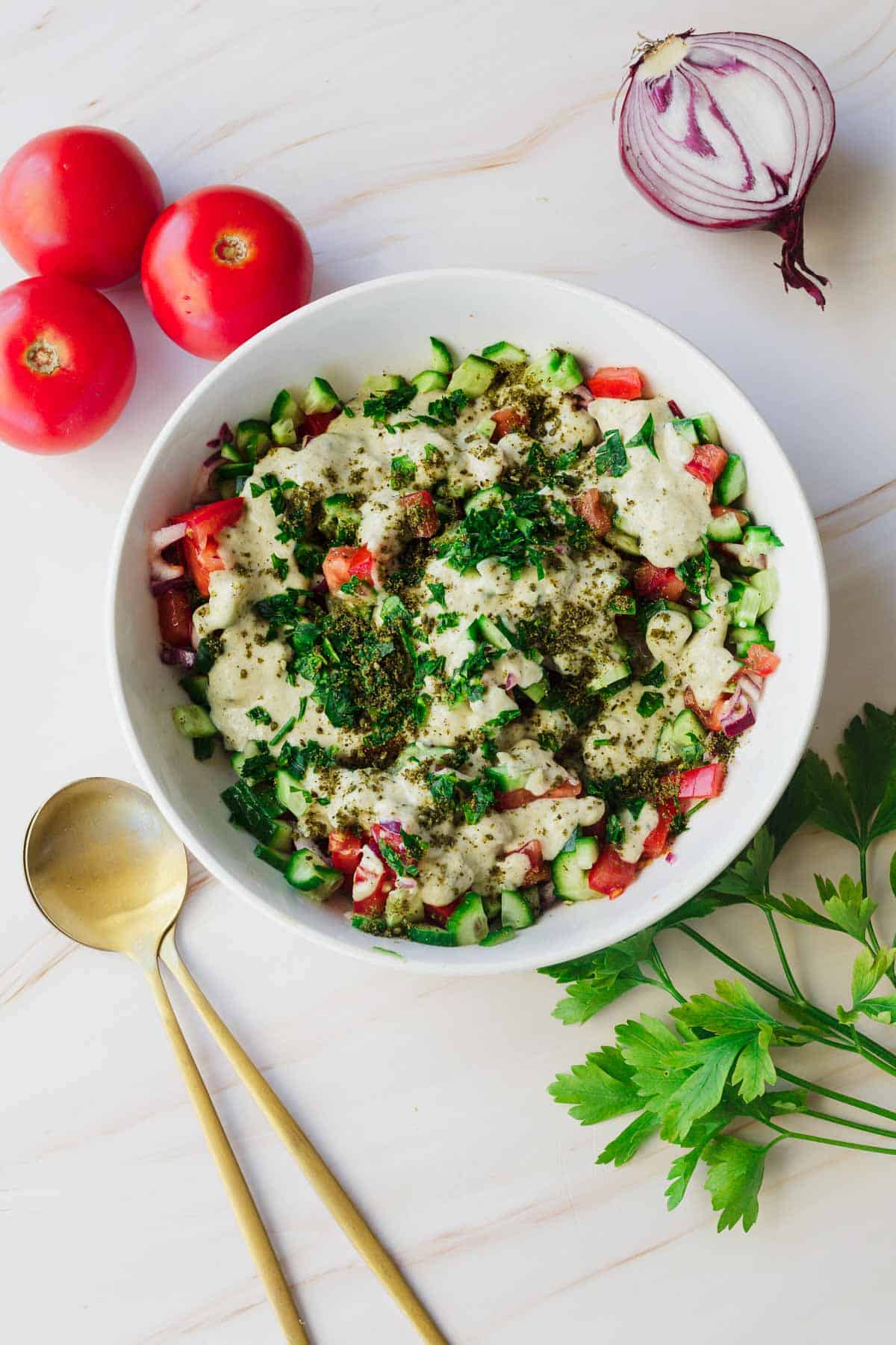 A bowl filled with a fresh Israeli chopped salad containing diced tomatoes, cucumbers, onions, and herbs, topped with a creamy dressing and sprinkled with seasonings. Surrounding the bowl are three whole tomatoes, half a red onion, a bunch of parsley, and two golden spoons.