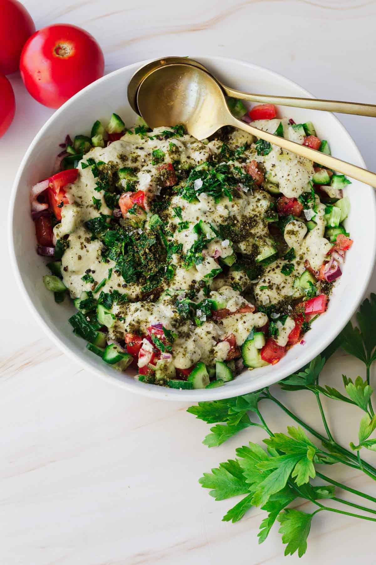 A bowl of creamy Israeli chopped salad featuring diced tomatoes, cucumbers, and red onions, topped with a dressing and sprinkled with herbs. Two gold-colored spoons rest in the bowl. Fresh tomatoes and parsley are placed around the bowl on a wooden surface.