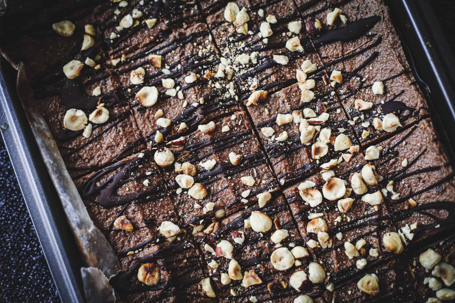 A tray of chocolate brownies topped with drizzled chocolate and chopped hazelnuts, cut into squares and still in the baking pan—perfect for trying a gluten free hazelnut bars recipe.
