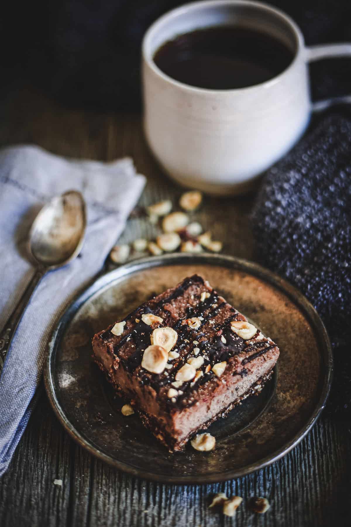 A square piece from a gluten free hazelnut bars recipe sits on a plate beside a cup of black coffee, a silver spoon, and a folded napkin on a dark wooden surface.