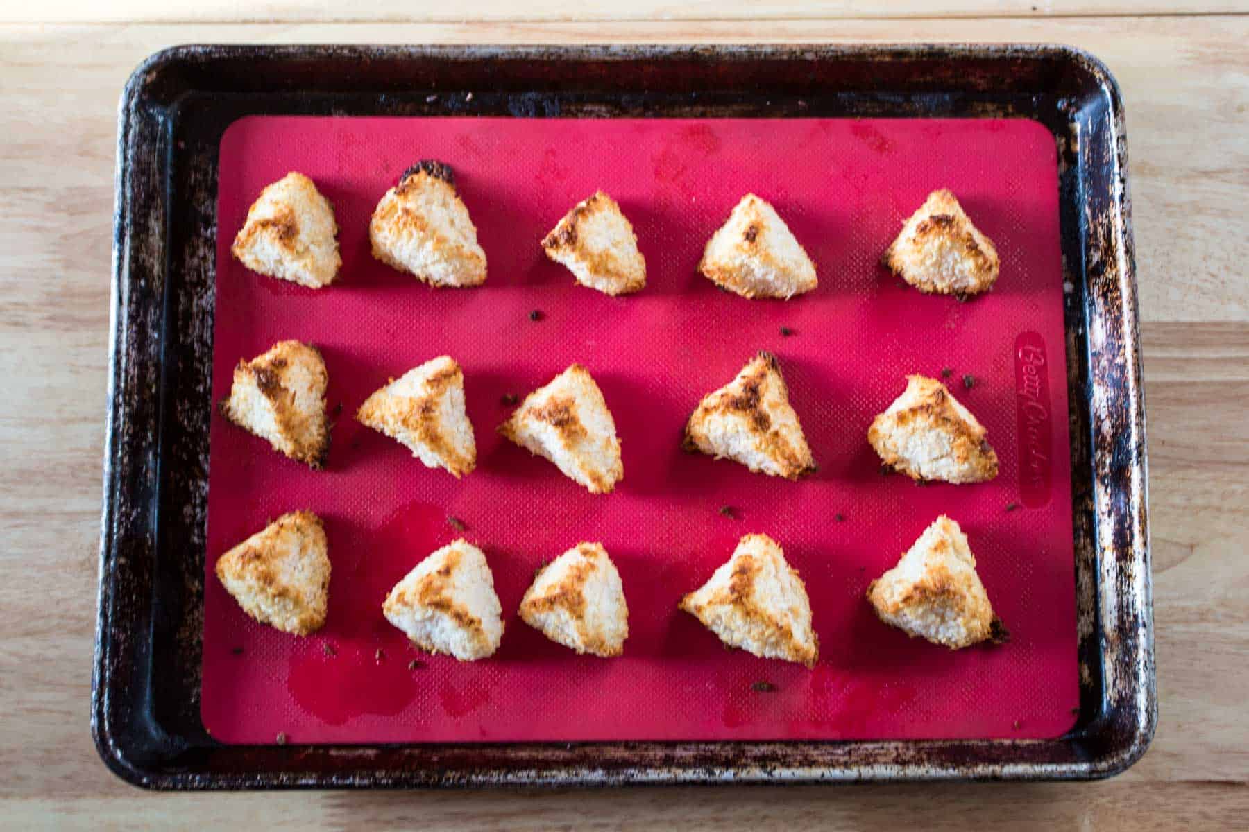 A baking tray with a red silicone mat holds 16 evenly spaced, triangular macaroon pyramids. These coconut delights are lightly toasted, arranged in four neat rows of four on the wooden surface.