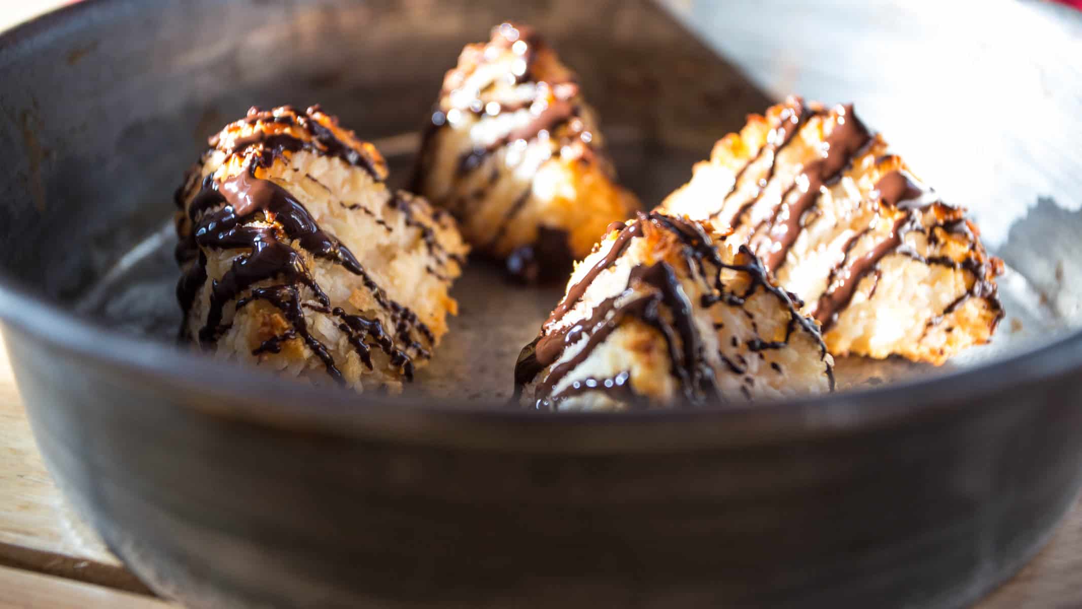 Four triangular coconut macaroon pyramids drizzled with chocolate sit on a ceramic plate. The plate rests on a wooden surface, where sunlight casts shadows across the scene.