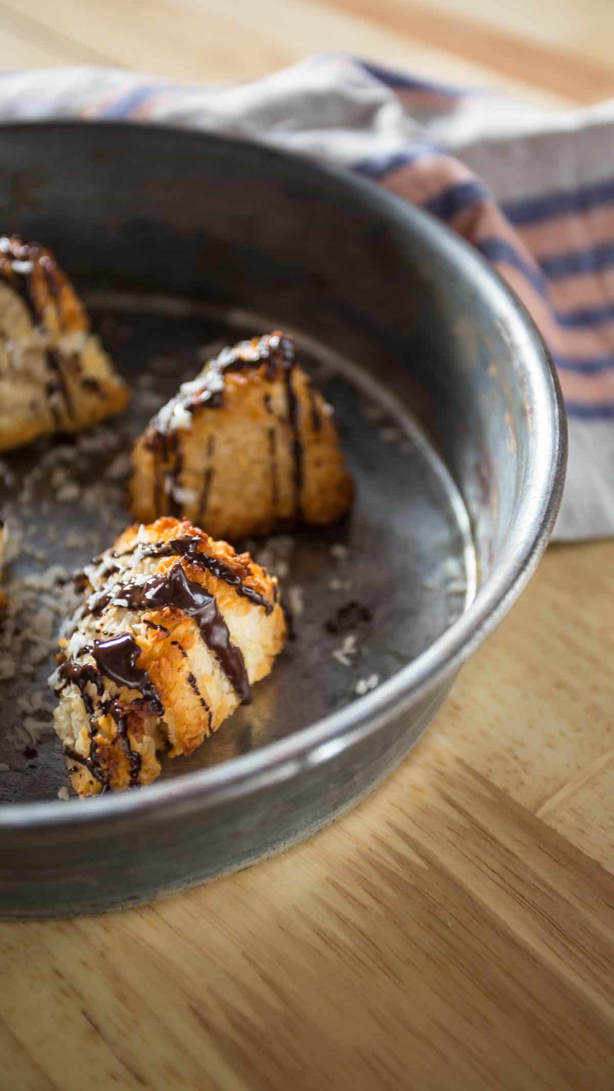 A round metal baking dish showcases several macaroon pyramids drizzled with chocolate. The dish rests on a wooden surface beside a striped cloth.