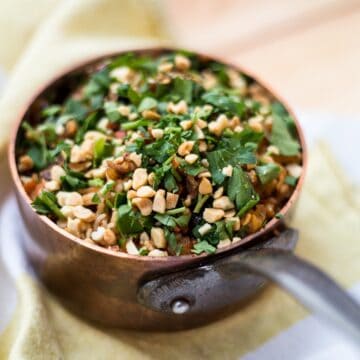 A metal pan filled with vegetarian pilaf, topped with chopped peanuts and fresh herbs, sits on a light-colored cloth.
