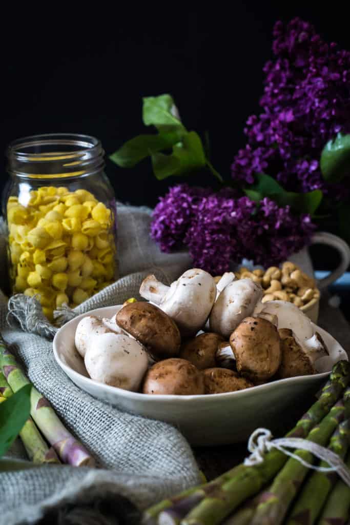 A white bowl filled with whole white and brown mushrooms sits on a table with fresh asparagus, a jar of yellow diced food, purple flowers, and some nuts in the background—perfect ingredients for a vegan pasta primavera.