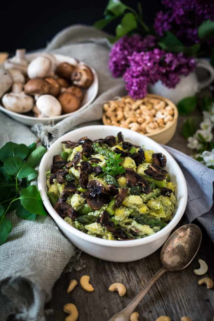 A white bowl filled with green salad and cooked mushrooms sits on a wooden table, surrounded by a spoon, raw cashews, whole mushrooms, and purple flowers—a scene reminiscent of a fresh vegan pasta primavera spread.