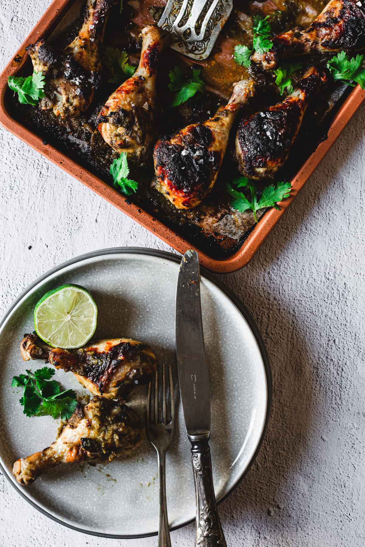 two roasted chicken legs with lime on a plate, fork and knife and tray of chicken drumsticks