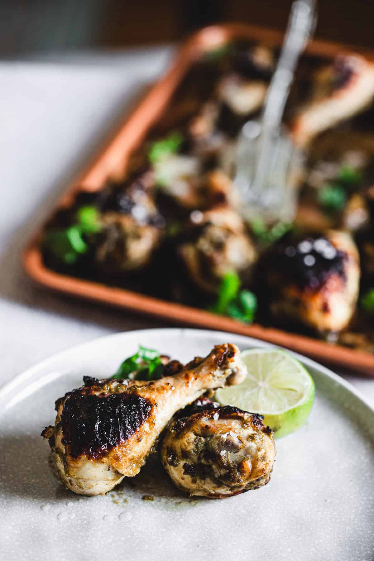 two roasted chicken legs with lime on a plate, tray of chicken drumsticks in background