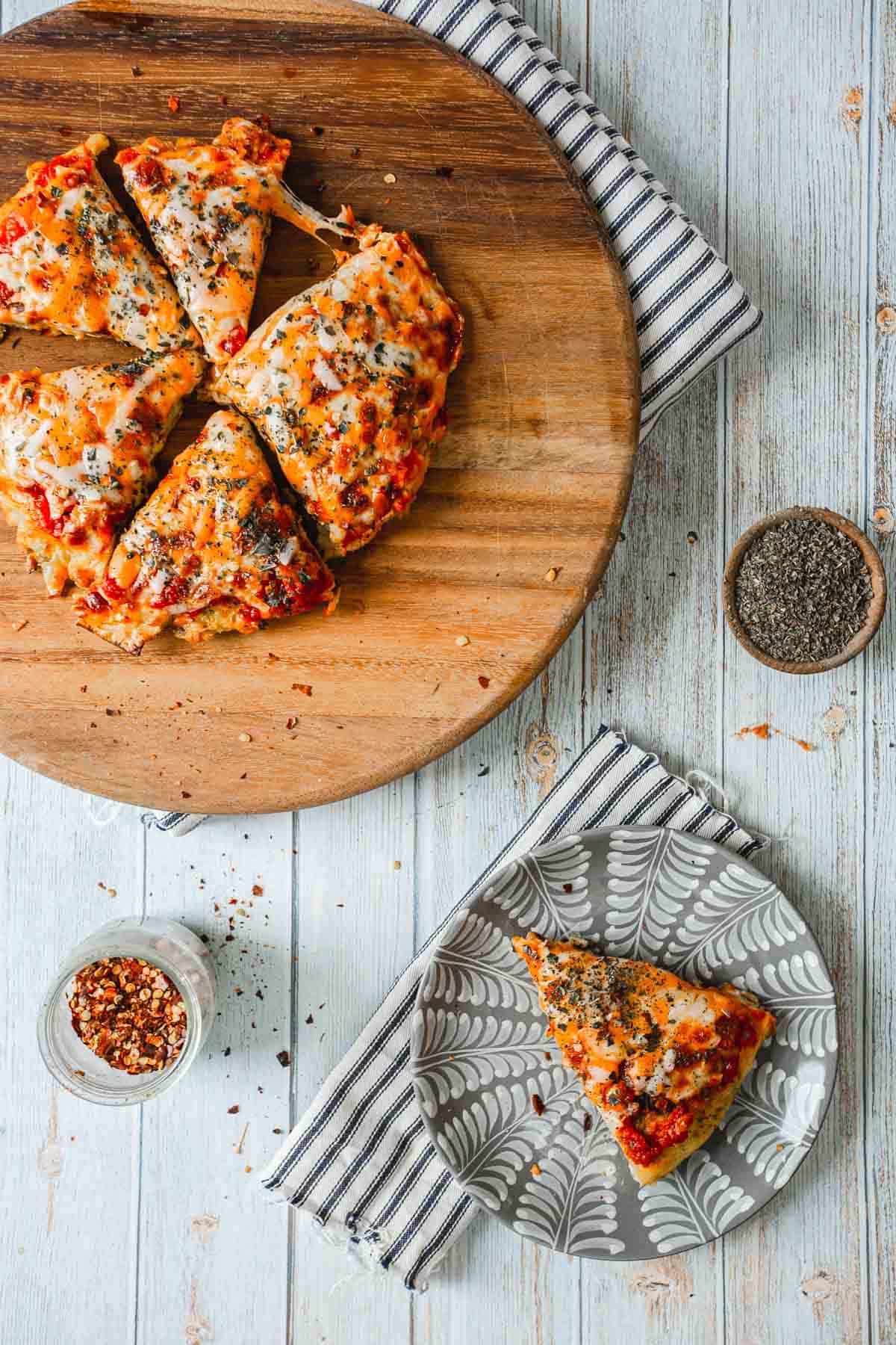 A round wooden board holds a sliced matzo brei pizza topped with cheese and herbs. One slice rests on a patterned plate beside the board, while a striped cloth lies beneath both. Small bowls of chili flakes and pepper are scattered across the light wooden surface.