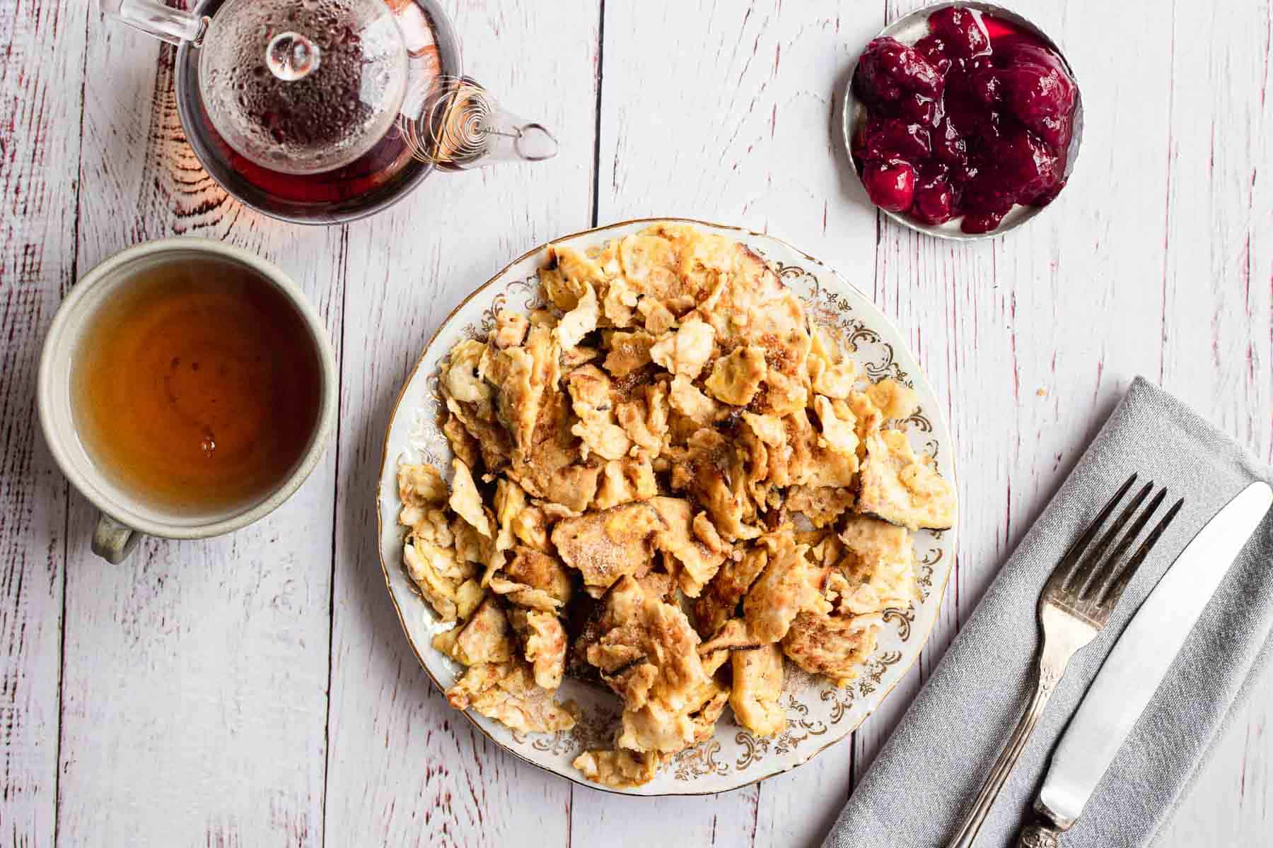 A wooden table is set with a plate of sweet matzo brie, a cup of tea, a teapot, a small dish of red jam, and a fork and knife on a gray napkin.