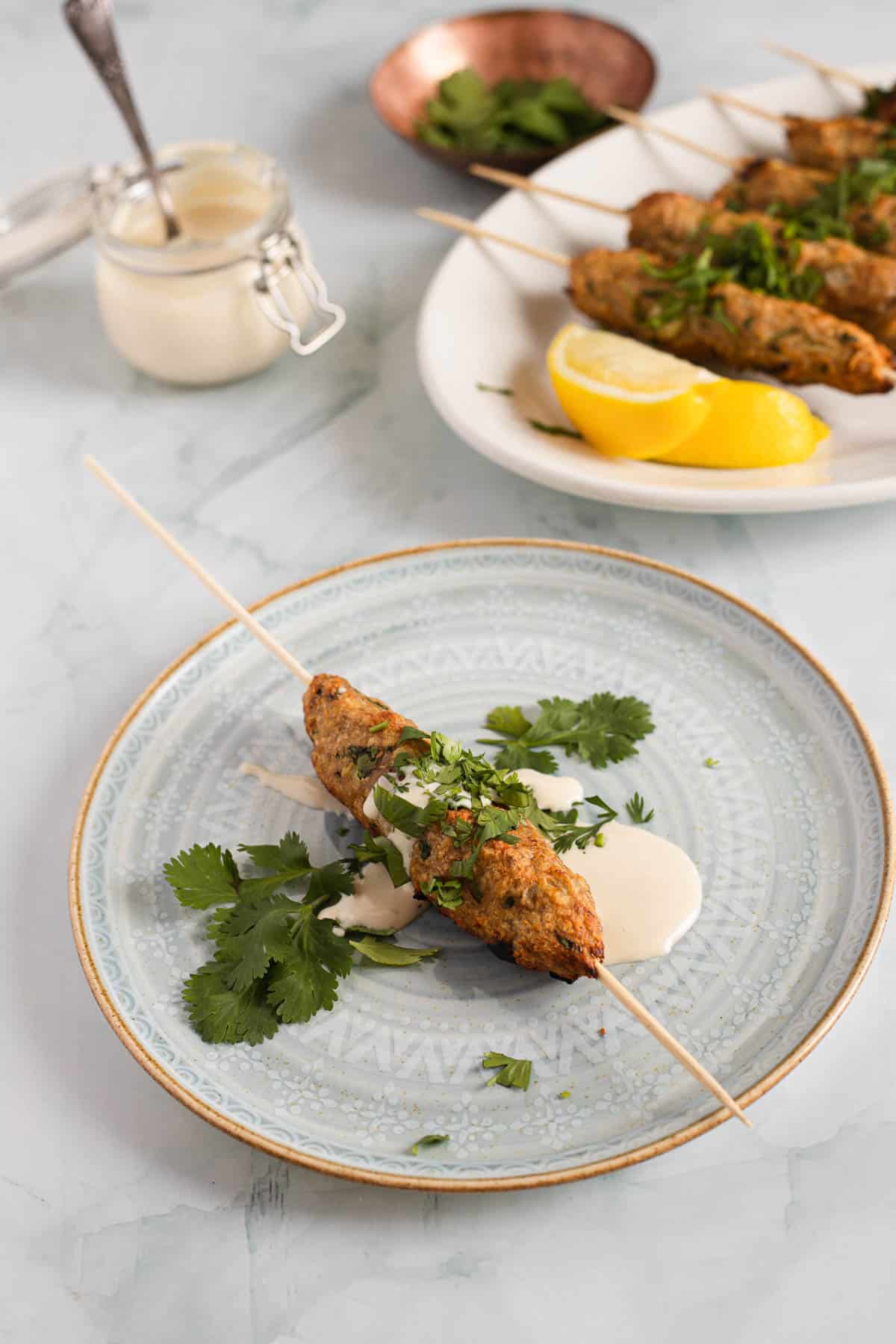 A whole kebab on a skewer on a plate with sauce and cilantro, more in the background on a plate.
