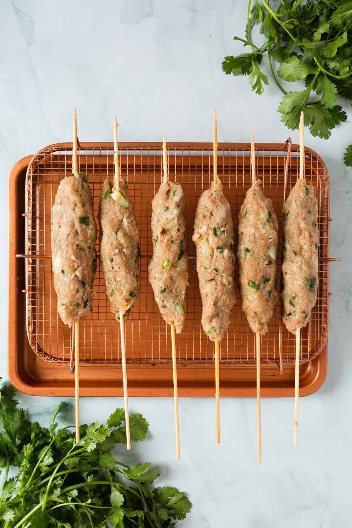 Six raw chicken kofta kebab skewers on a baking sheet with cilantro.