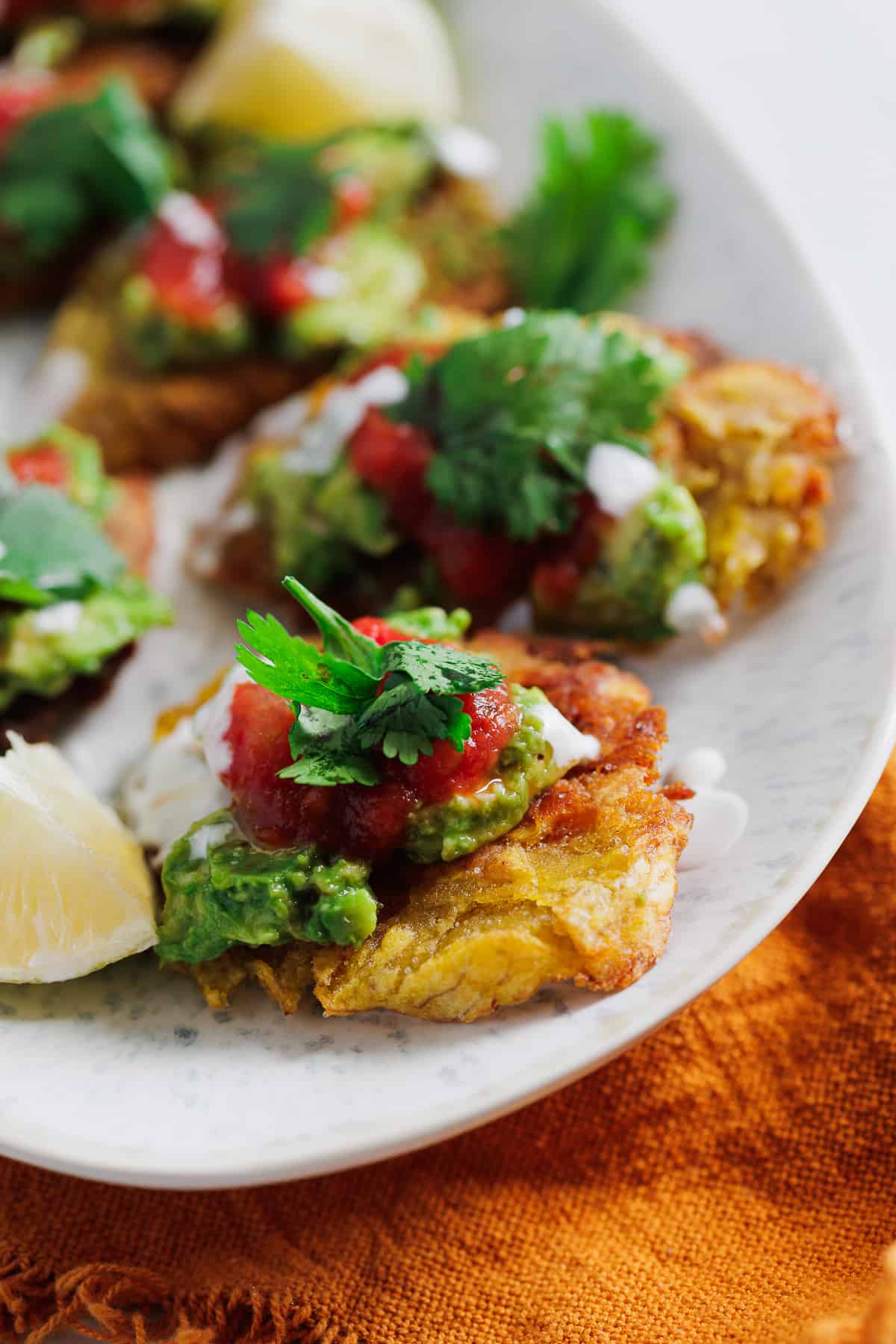 A plate of traditional fried green plantains topped with guacamole, salsa, and a dollop of cream garnished with cilantro, accompanied by lemon wedges on the side.