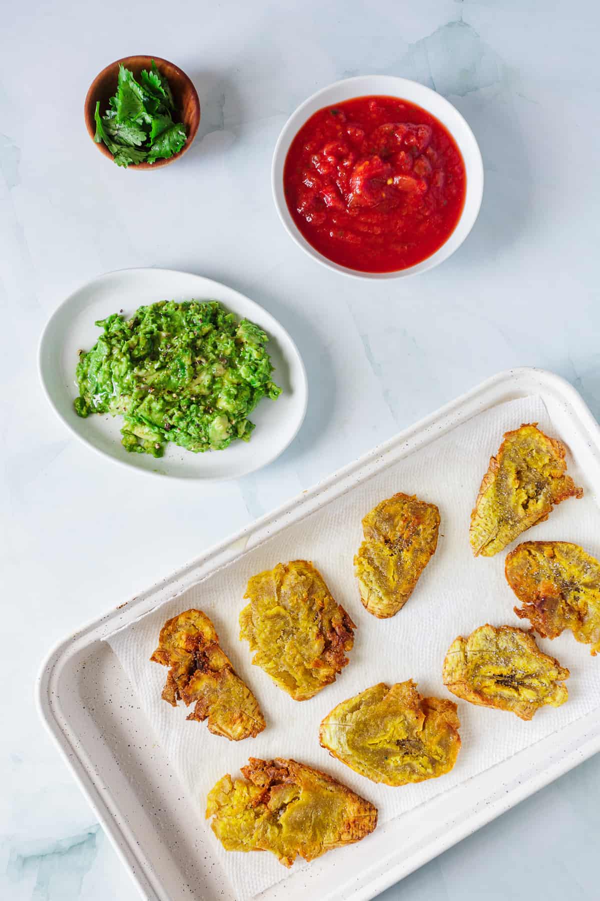 A tray of traditional Colombian Patacones, served with two bowls of dip, one red and one green, on a marble surface.