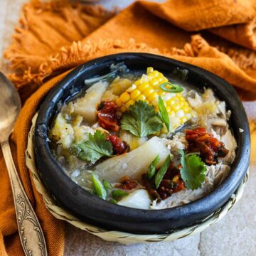 A traditional stone bowl containing a chicken sancocho stew with corn, meats, and garnished with fresh herbs, presented on a table with a rustic orange napkin and a wooden spoon.