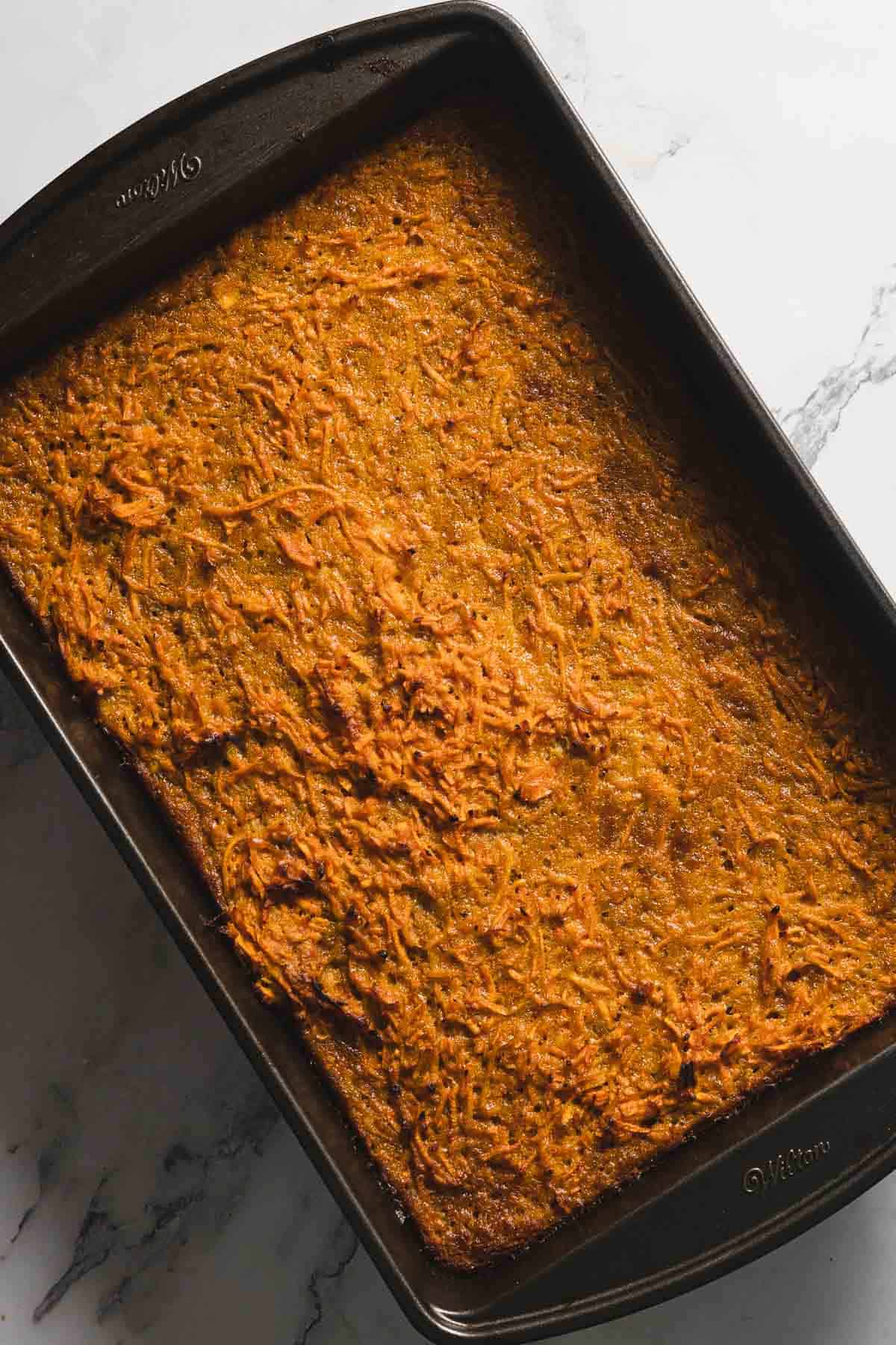 A rectangular baking dish containing a freshly baked carrot cake placed on a marble countertop. The surface is golden brown and slightly uneven, showing grated bits of carrot.