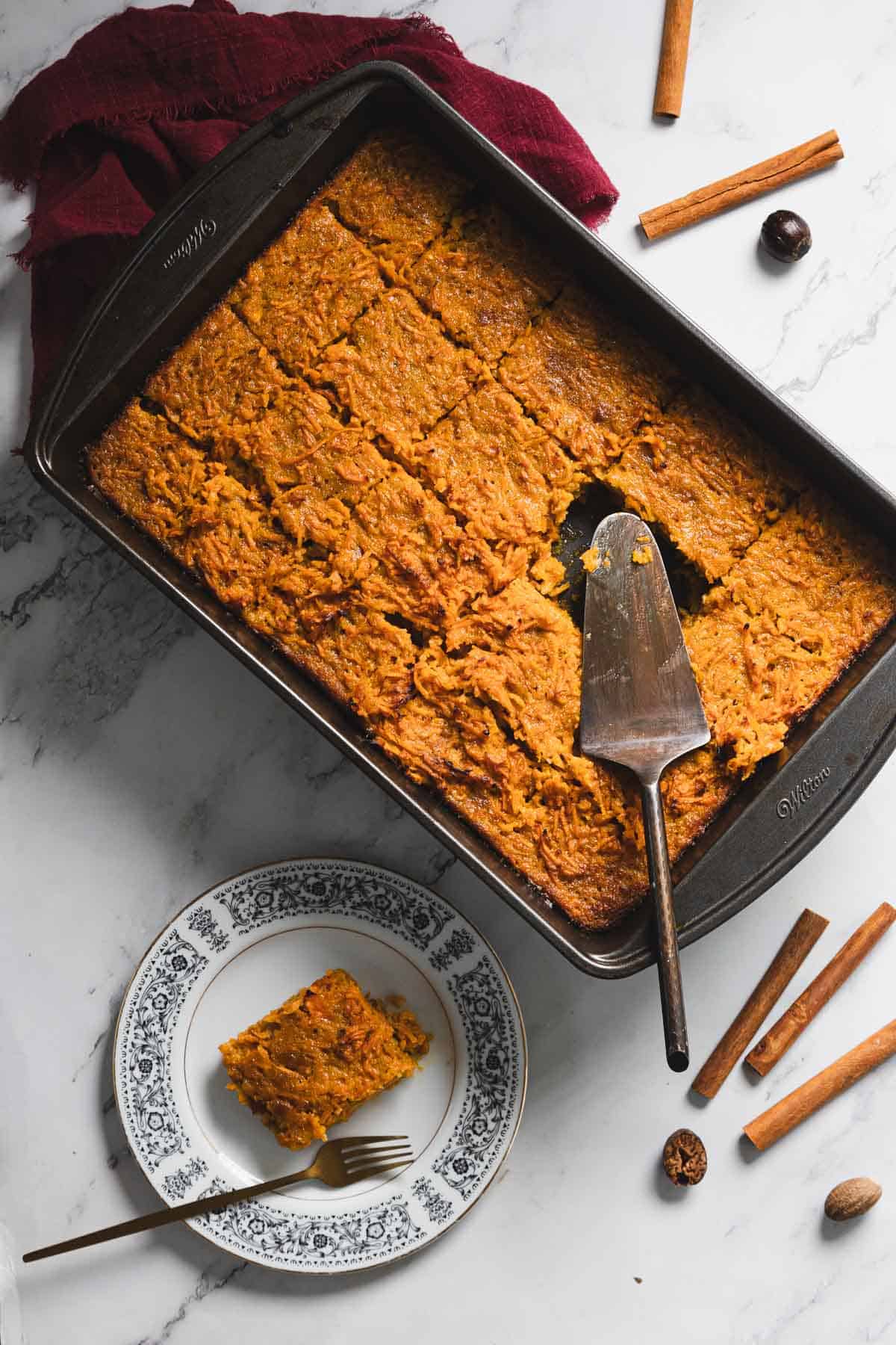 A rectangular baking dish filled with sliced baked pumpkin treat rests on a marble countertop. A slice is served on a decorative plate beside the dish. A serving spatula lies on the dish. Cinnamon sticks and whole nutmeg are scattered around.
