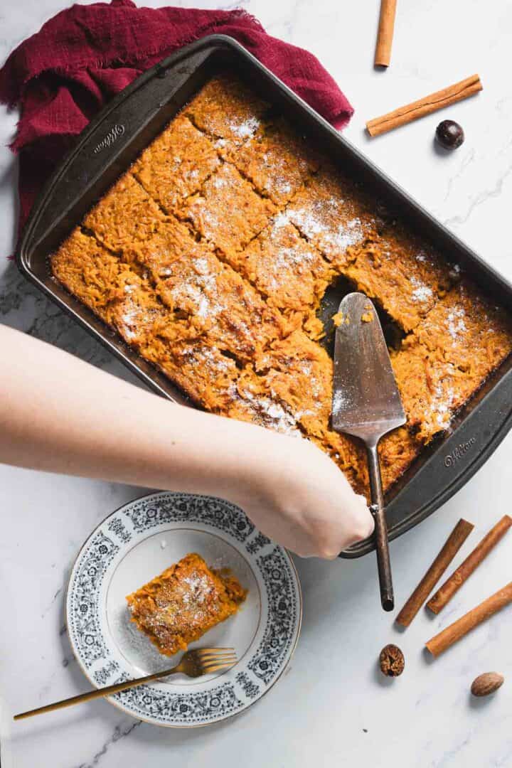 A rectangular baking dish filled with sliced baked pumpkin treat rests on a marble countertop. A slice is served on a decorative plate beside the dish. A serving spatula lies on the dish. Cinnamon sticks and whole nutmeg are scattered around.