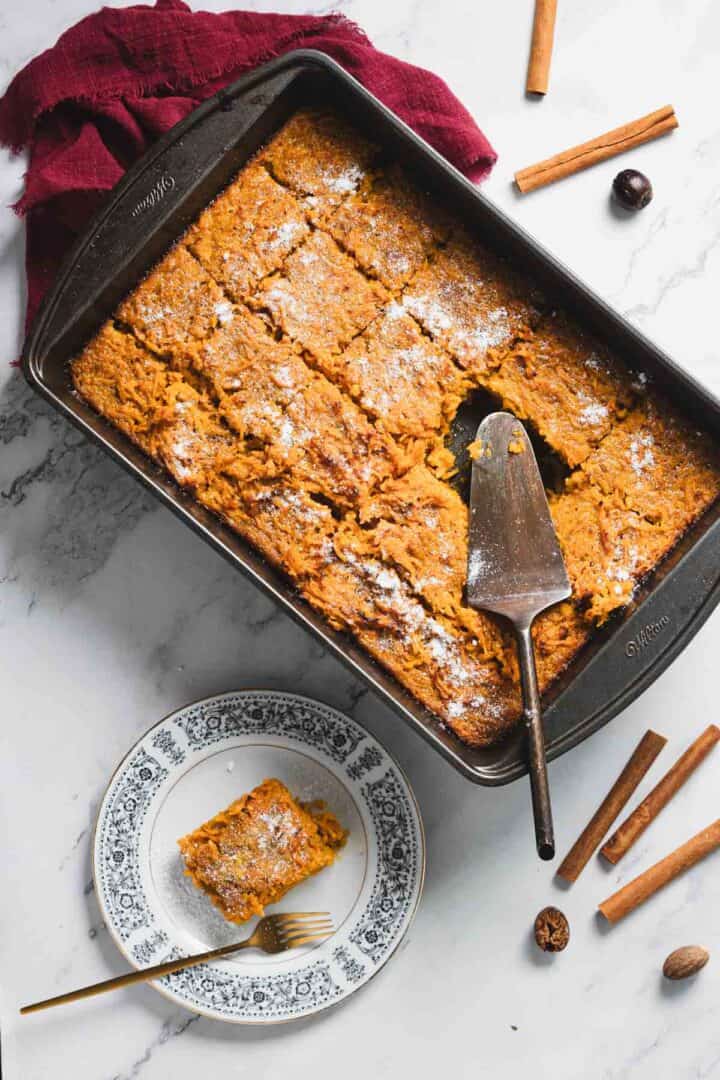A rectangular baking dish filled with sliced baked pumpkin treat rests on a marble countertop. A slice is served on a decorative plate beside the dish. A serving spatula lies on the dish. Cinnamon sticks and whole nutmeg are scattered around.