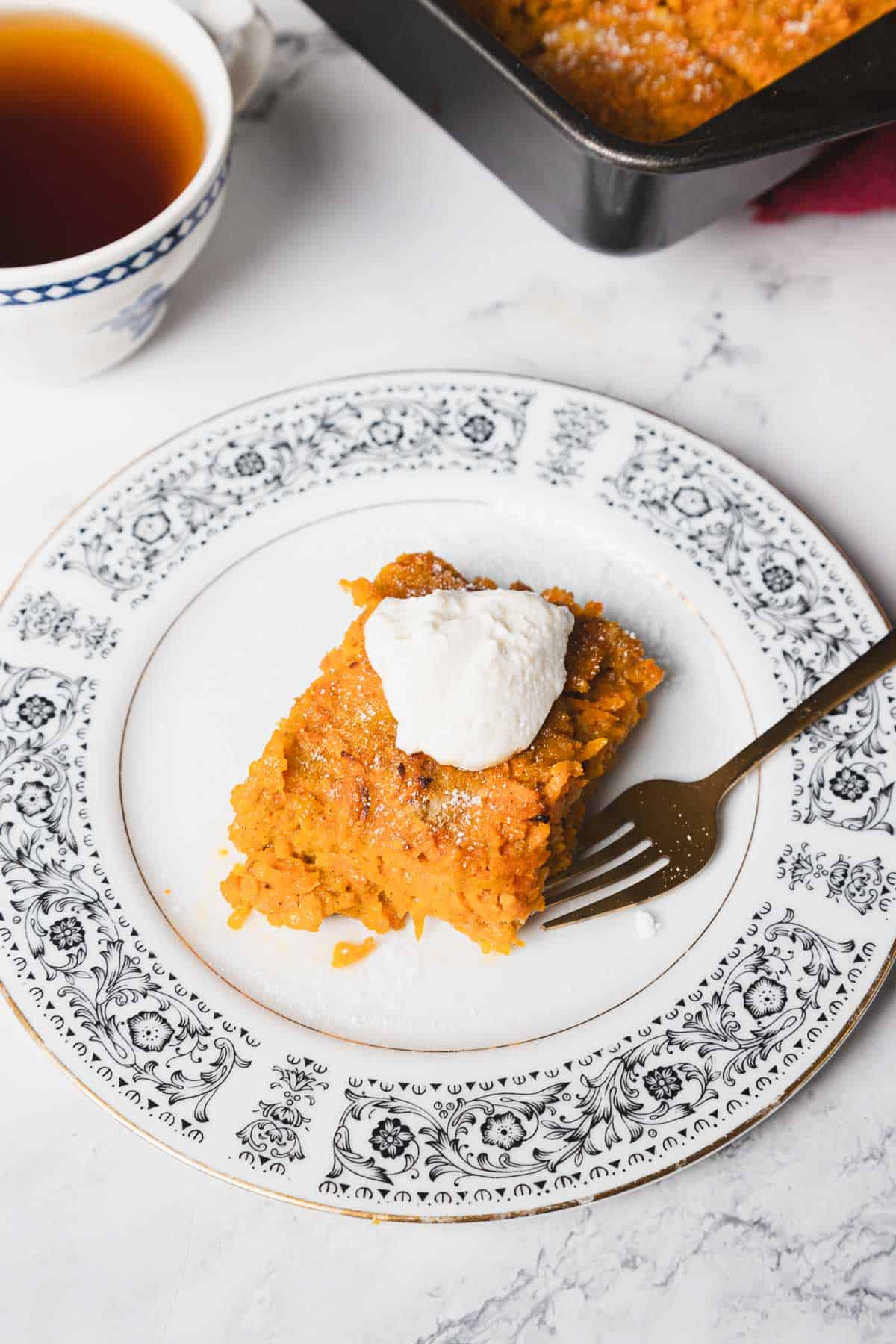 A carrot kugel slice topped with a dollop of cream sits on an ornate patterned white plate with a fork beside it. The dessert has a yellowish-orange color and is placed on a white marble surface. A black baking tray with more dessert is partially visible in the background.