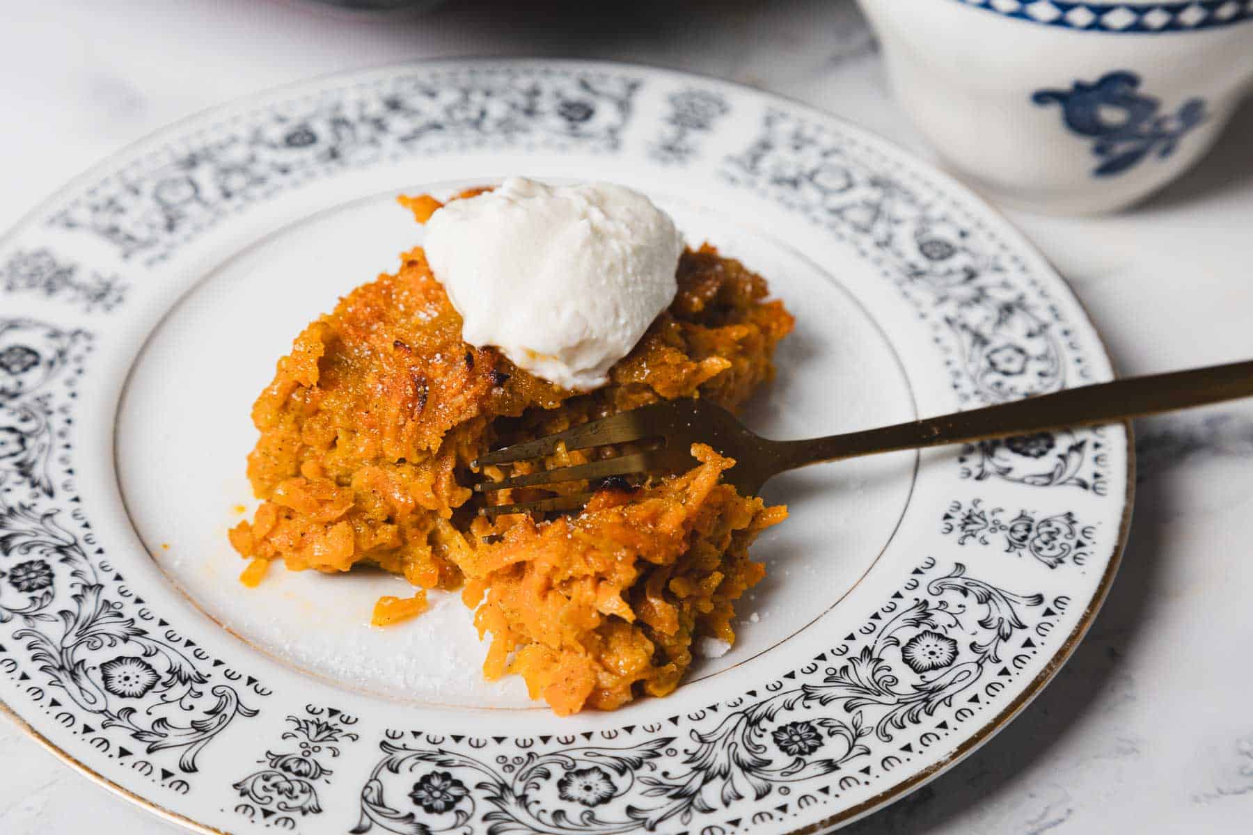 A carrot kugel slice topped with a dollop of cream sits on an ornate patterned white plate with a fork beside it. The dessert has a yellowish-orange color and is placed on a white marble surface. A black baking tray with more dessert is partially visible in the background.