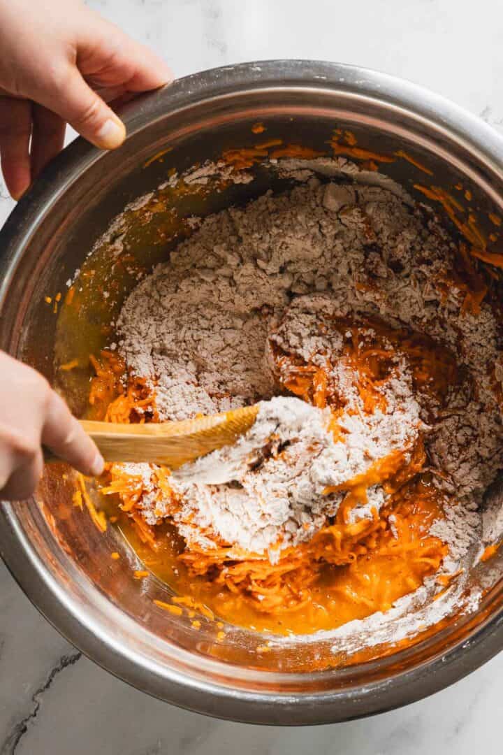 A close-up image of a large metal mixing bowl with shredded carrots, eggs, and brown sugar being mixed with a wooden spoon. Two hands are visible, holding the bowl and the spoon. The mixing bowl is placed on a white marble countertop.