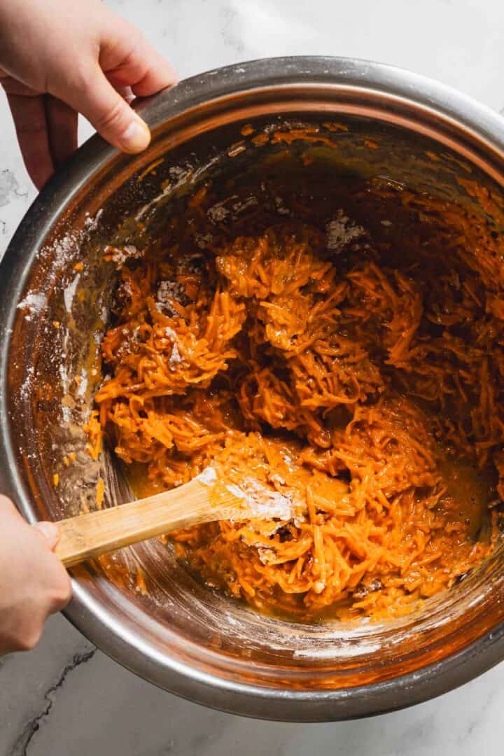 A close-up image of a large metal mixing bowl with shredded carrots, eggs, and brown sugar being mixed with a wooden spoon. Two hands are visible, holding the bowl and the spoon. The mixing bowl is placed on a white marble countertop.