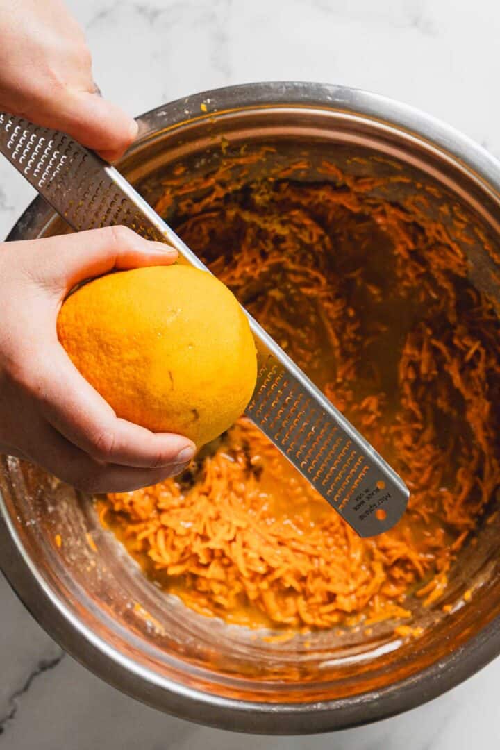 A person is zesting an orange over a stainless steel mixing bowl filled with shredded orange-colored ingredients. The person is using a grater to collect the orange zest. The background is a marble countertop.
