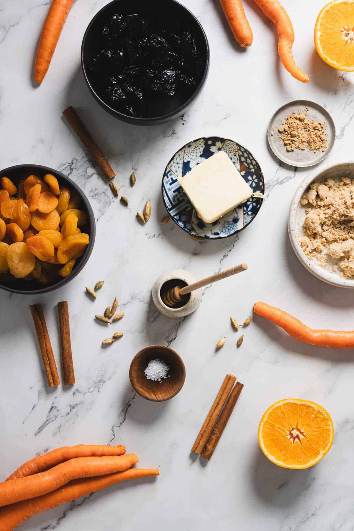 A top-down view of various ingredients on a white marble surface. Visible items include carrots, dried apricots, prunes, a stick of butter on a small plate, spices, brown sugar, half an orange, cinnamon sticks, and a honey dipper in a small bowl.