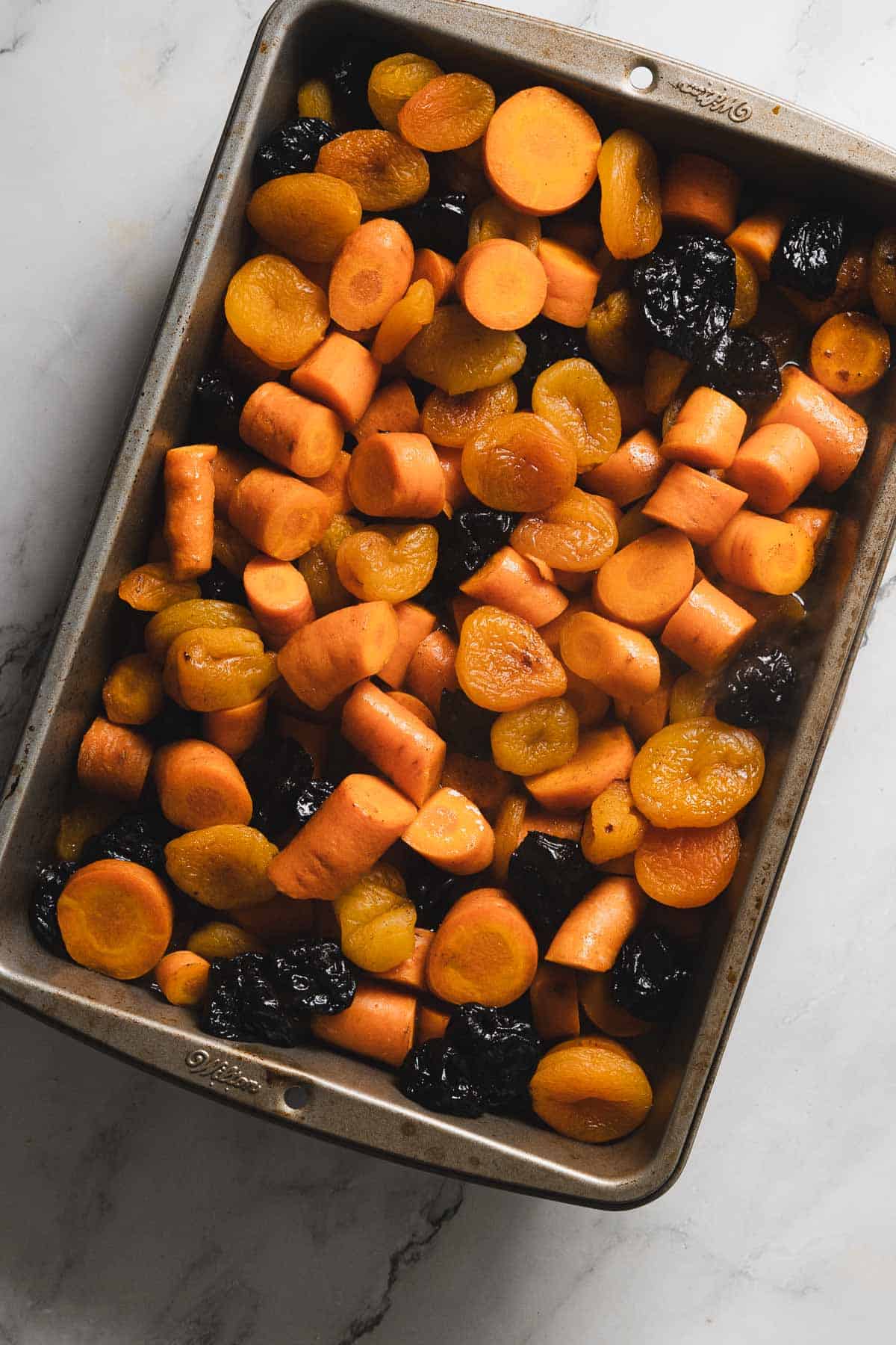 A baking tray filled with roasted root vegetables, including sliced carrots, apricots, and prunes, placed on a white marbled surface.