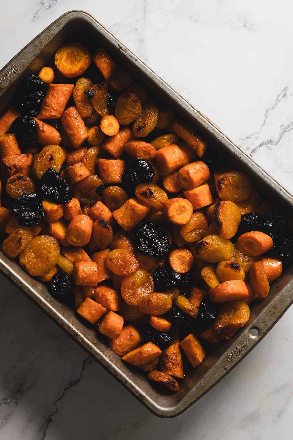A metal baking dish filled with roasted vegetables, including chopped sweet potatoes, carrots, and prunes, placed on a marble surface. The vegetables are browned and appear well-cooked.