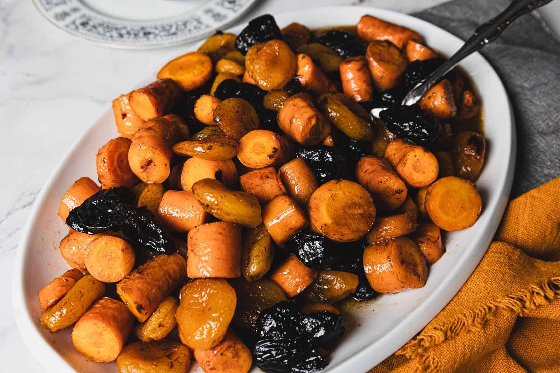 A white oval dish holds a serving of tzimmes, a traditional Jewish stew, made with chunks of glazed carrots, sweet potatoes, and prunes. The dish is placed on a white and gray marble surface next to a yellow cloth napkin and a gray towel.