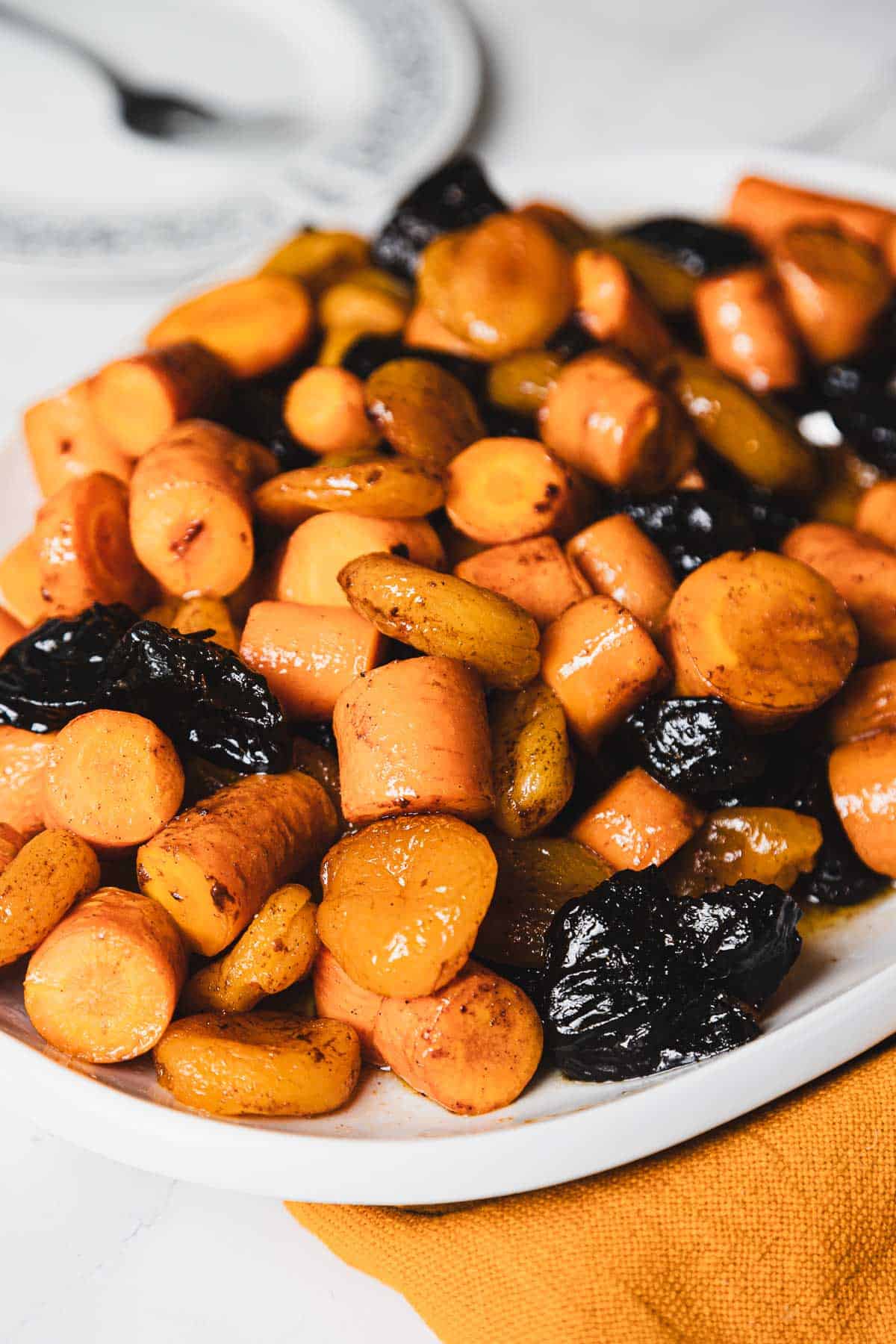 A close-up of a white plate filled with glazed carrot coins and prunes. The food appears to be seasoned and cooked, laid out on an orange cloth placed on a white surface. A fork and a partially visible plate are in the background.