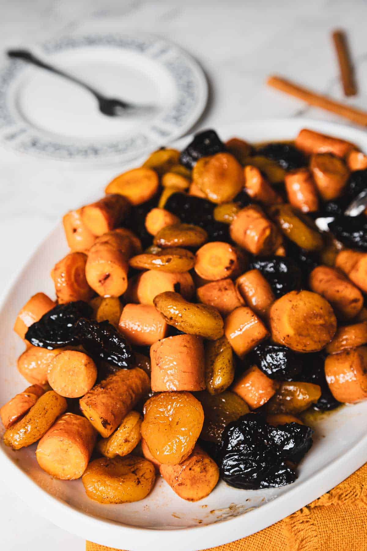 A white plate is filled with tzimmes, a traditional Jewish dish made of glazed carrots, sweet potatoes, prunes, and apricots. A fork rests on a dish in the background, placed on a white tablecloth. Two cinnamon sticks lie beside the plate.