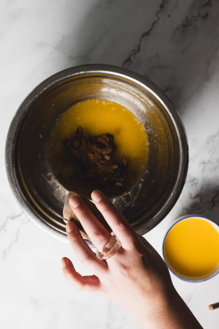 A hand holding brown sugar or a spice is about to add it to a bowl with melted butter. A small white and blue cup containing yellow liquid is placed near the bowl on a marble countertop.