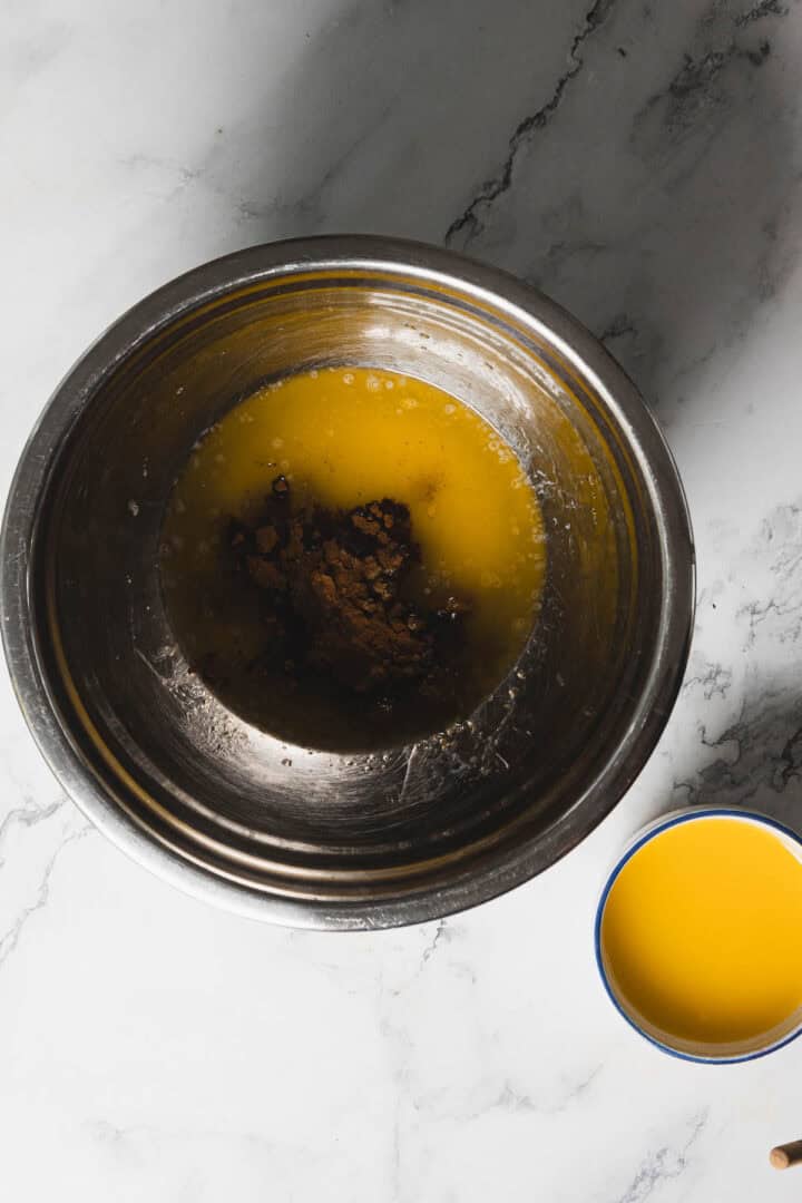 A stainless steel mixing bowl contains melted butter and dark brown sugar. Next to the bowl is a cup filled with orange liquid on a white marble surface.