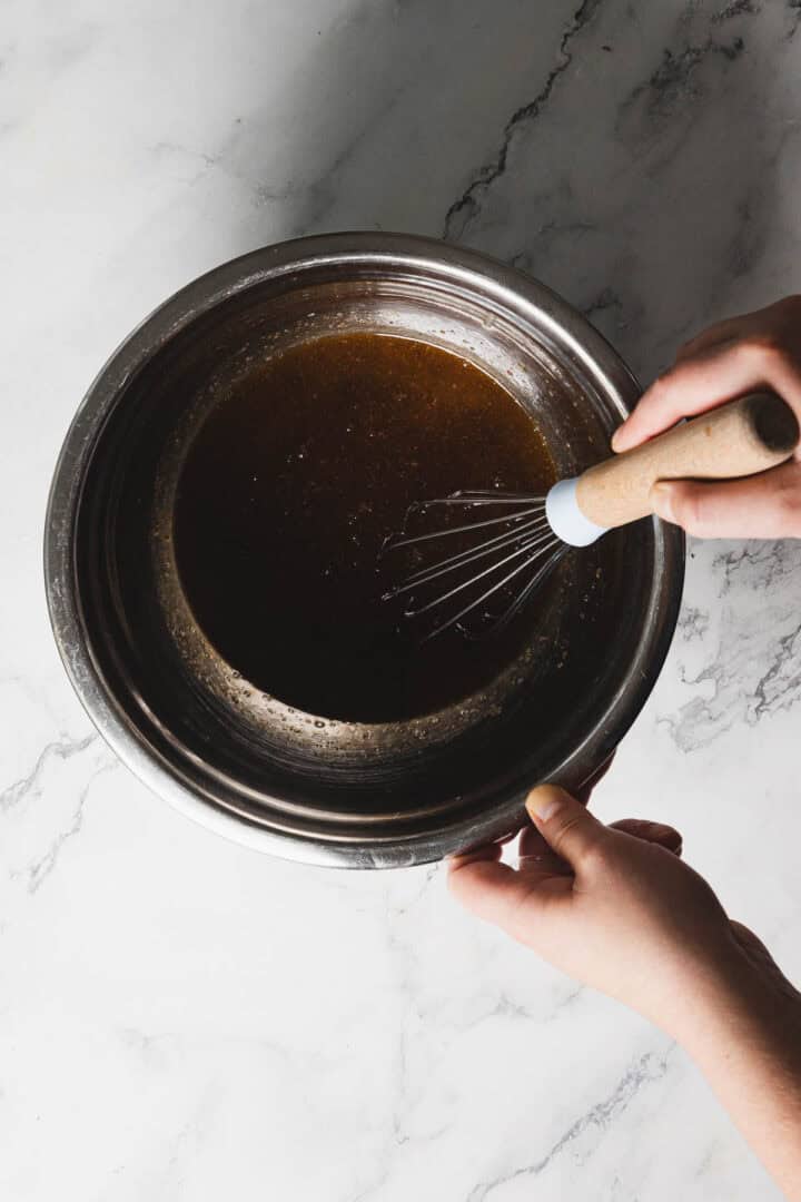 A person stirring a dark brown liquid in a stainless steel bowl using a whisk with a wooden handle. The bowl is on a white marble surface. Only the person's hands are visible.