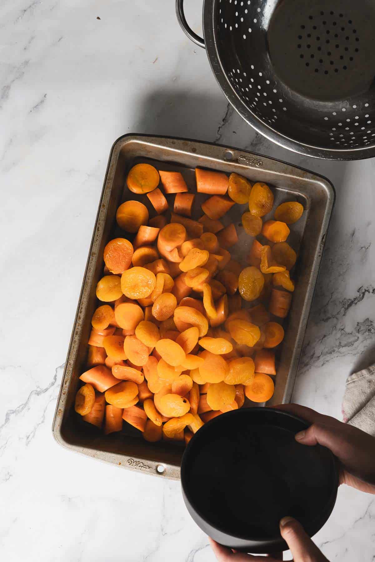 A baking tray filled with sliced orange vegetables, likely carrots, sits on a marble countertop. A colander is positioned nearby in the upper right corner. Hands are seen holding a black bowl in the lower right section of the image.